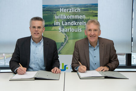 Zwei Männer sitzen an einem Tisch und unterzeichnen Dokumente. Links der Oberbürgermeister der Kreisstadt Merzig, rechts der Landrat des Landkreises Saarlouis.