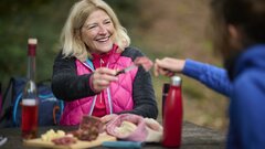 Wanderin genießt eine Picknickpause im Wald und reicht einer Begleitperson ein Stück Wurst.