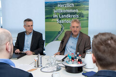 Mehrere Personen sitzen an einem Konferenztisch im Gespräch. Links der Oberbürgermeister der Kreisstadt Merzig, rechts der Landrat des Landkreises Saarlouis.